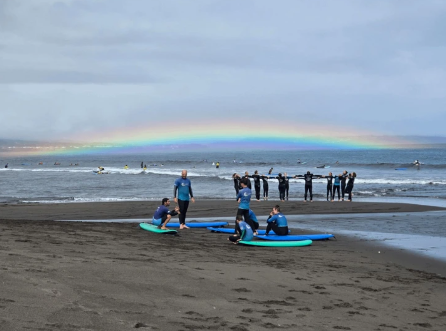 Surferi na čiernom piesku, dúha nad oceánom, výcviková lekcia surfovania, plážová atmosféra.