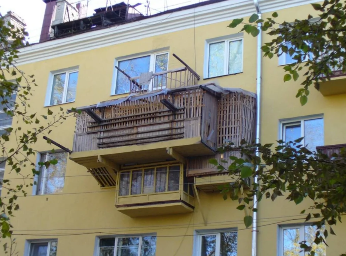 Pochybná konštrukcia balkóna z dreva a kovu na žltej budove s dvomi oknami a okolím so stromami.