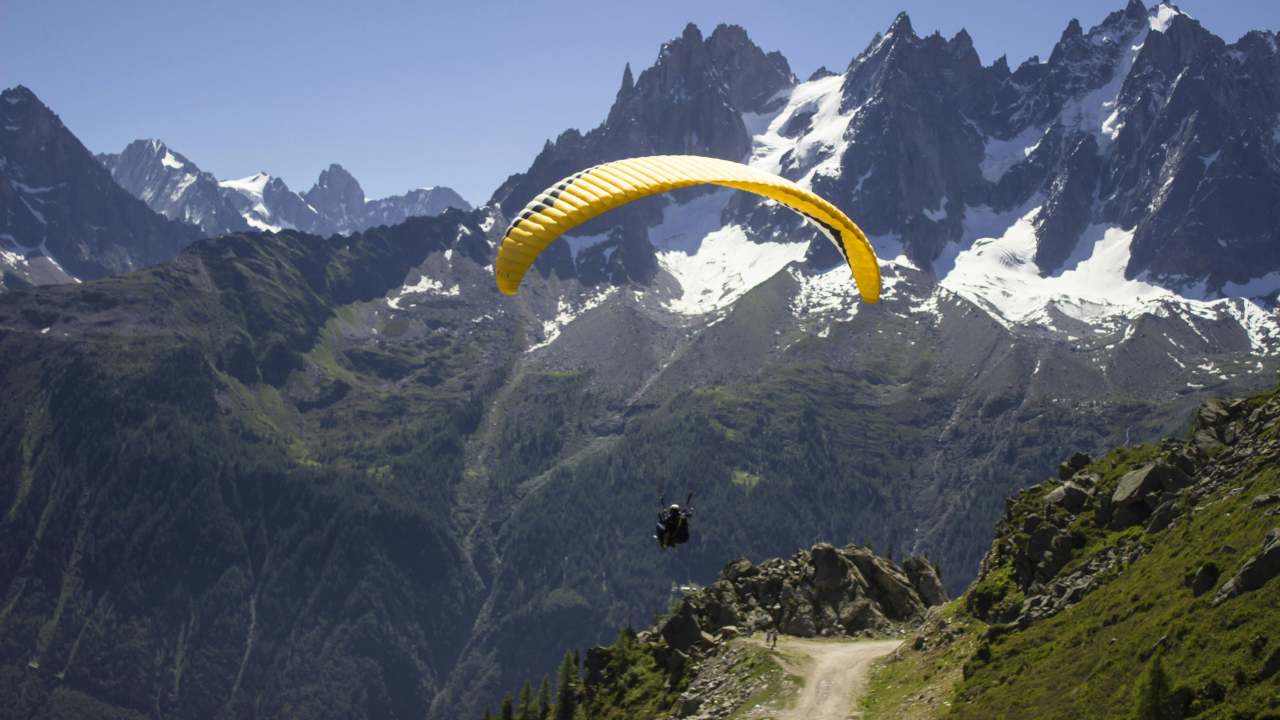 Paraglider s žltým krídlom letiaci nad horskou krajinou so zasneženými vrcholmi.