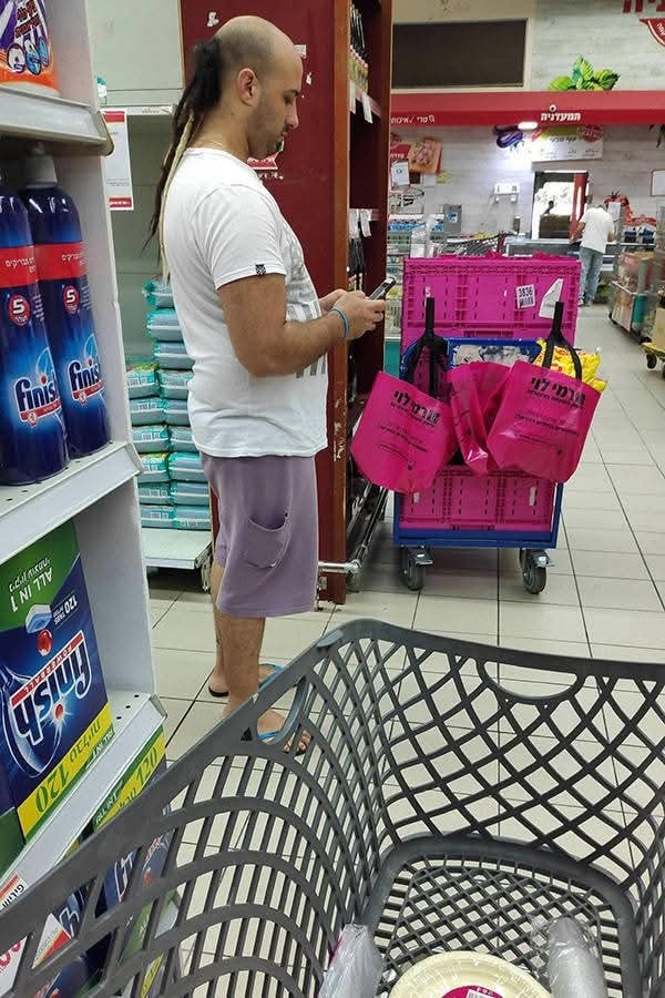 Osoba s neobvyklým účesom nakupuje v supermarkete, vloží produkty do košíka a kontroluje mobilný telefón.