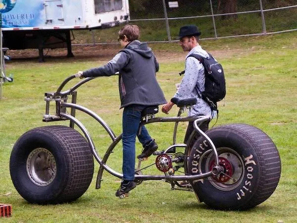 Dvaja ľudia skúšajú jazdu na obrím bicykli s veľkými pneumatikami na trávnatom povrchu.