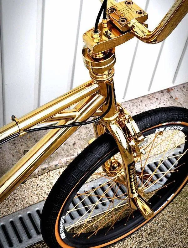 Zlatý bicykel so žiarivým rámom, čiernymi pneumatikami a zlatými lúčmi na ceste pri bielej stene.