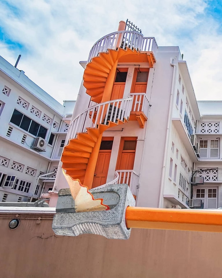 Oranžové točité schody spojené s ceruzkou a strúhadlom pred modernou budovou pod modrou oblohou.