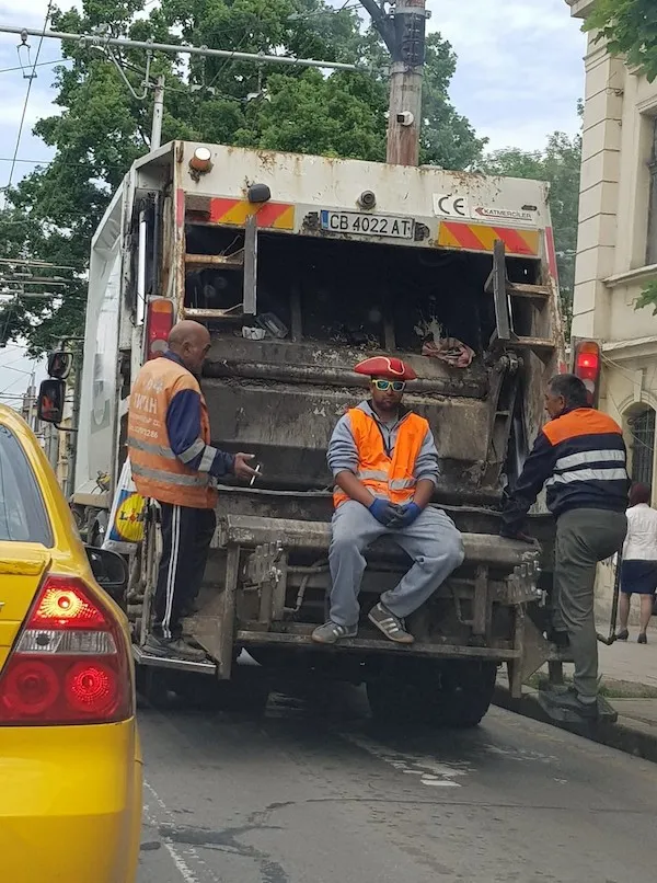 Muž s červeným klobúkom sedí na smetiarskom aute, oblečený v oranžovej veste, zatiaľ čo auto stojí na ulici.