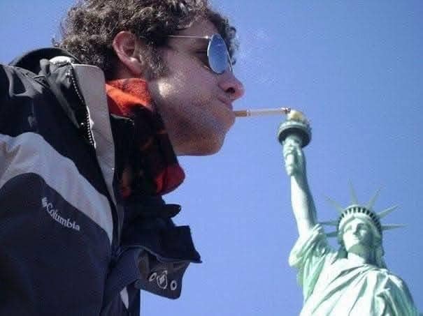 Muž fajčiaci cigaretu, ktorá vyzerá, akoby bola zapálená od pochodne Sochy slobody, vtipná perspektíva.