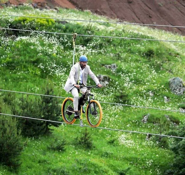Muž v obleku jazdiaci na bicykli po lane nad zelenou lúkou, nosí prilbu pre bezpečnosť.