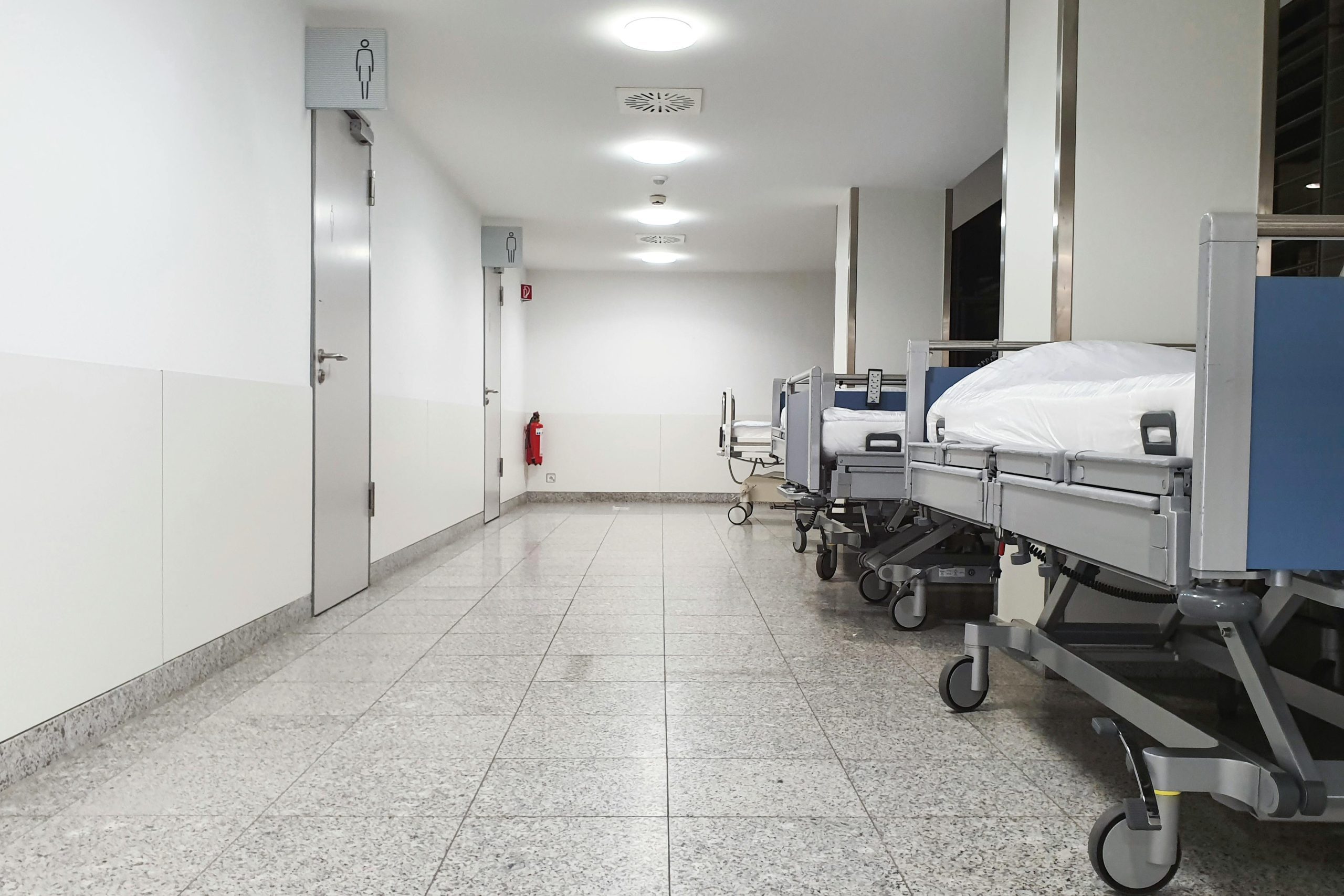 Nemocničná chodba s prázdnymi lôžkami a dverami toaliet v sterilnom prostredí.