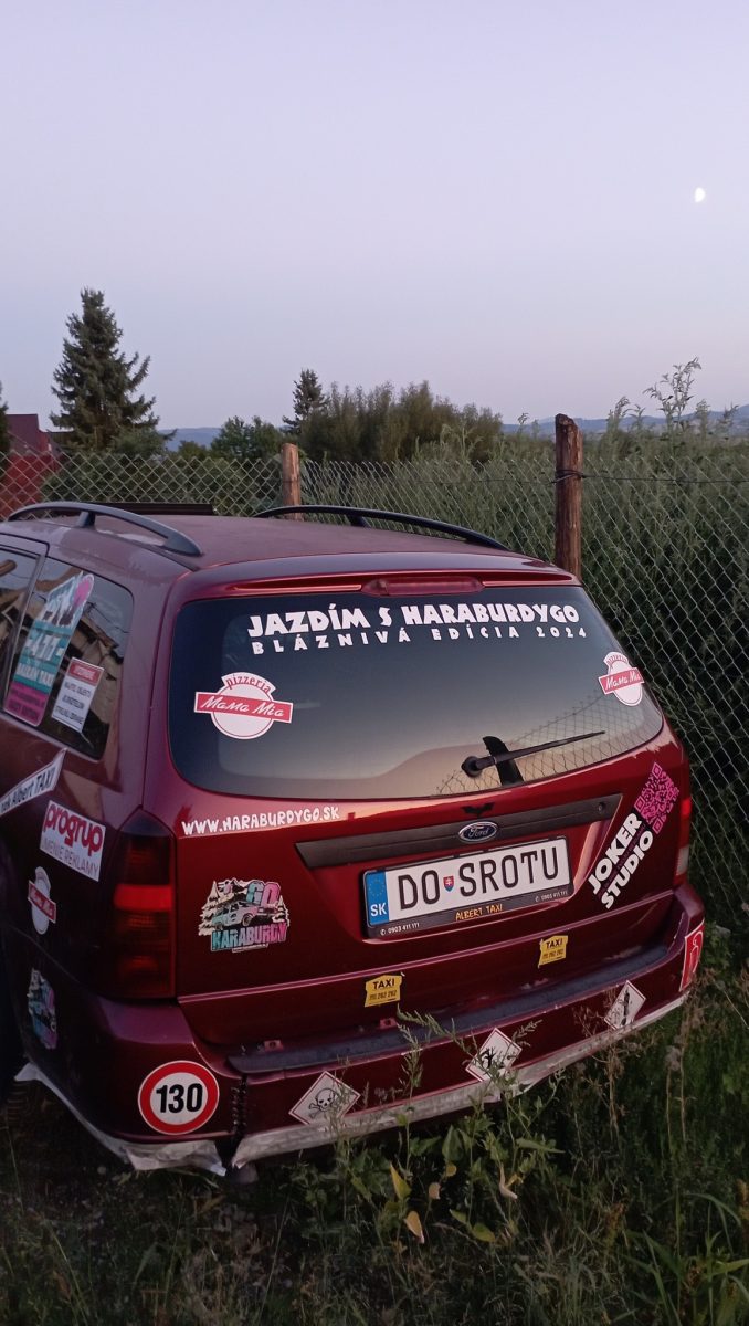 Auto s reklamnými nálepkami a poznávacou značkou DO SROTU zaparkované pri plote na vidieku.