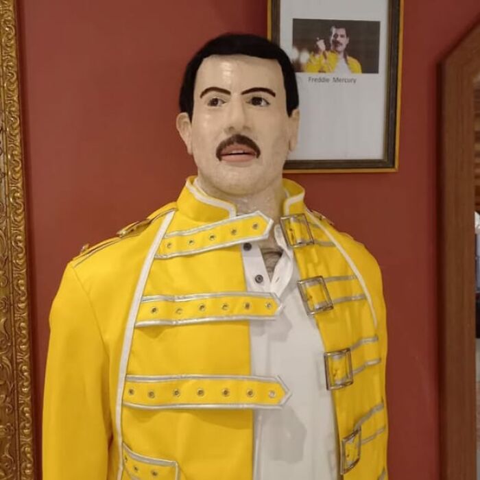 Socha muža v žltom kostýme s portrétom na stene v pozadí.