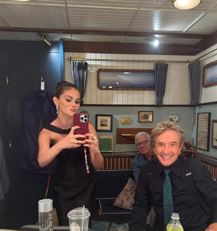 Žena v elegantných šatách si robí selfie v miestnosti so starším pánom a ďalším mužom v pozadí.