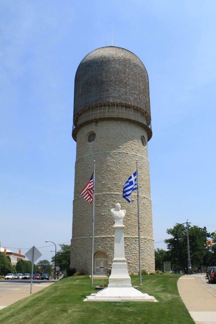 Kamenná veža s kupolou, pred ňou busty a americké a grécke vlajky, modré nebo.