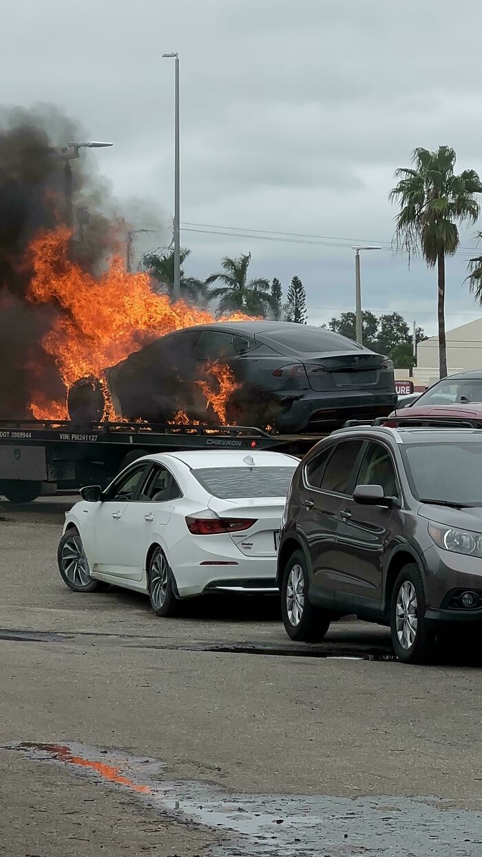 Auto v plameňoch na vozíku, v pozadí palmy a ďalšie autá na parkovisku.