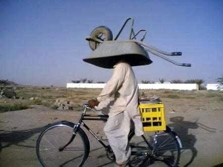 Muž na bicykli nesie na hlave fúrik, za ktorým je upevnená debna. Scéna na vidieku.