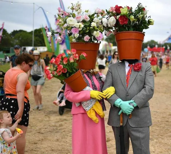Dve osoby v kostýmoch kvetináčov na festivale, oblečené v elegantných šatách a rukaviciach, s kvetmi na hlavách.