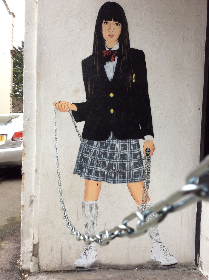 Graffiti dievča so školskou uniformou drží reťaz, ktorá visí z obrazu na stene.