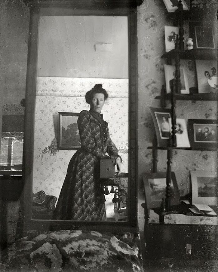 Žena s fotoaparátom v zrkadle, viktoriánska éra, čiernobiela fotka, historický interiér so starými fotografiami.