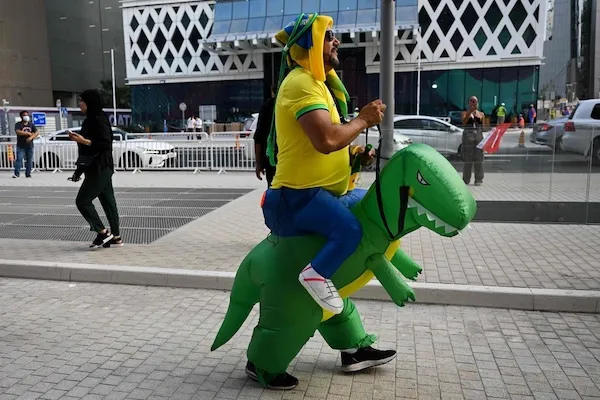 Muž v kostýme jazdí na nafukovacom dinosaurovi na ulici, okoloidúci v pozadí, moderné budovy na pozadí.