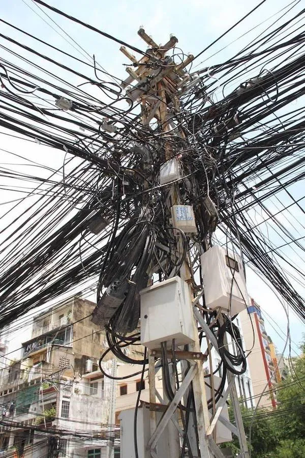 Zamotaný elektrický stĺp s množstvom prepletených káblov v rušnej mestskej oblasti.