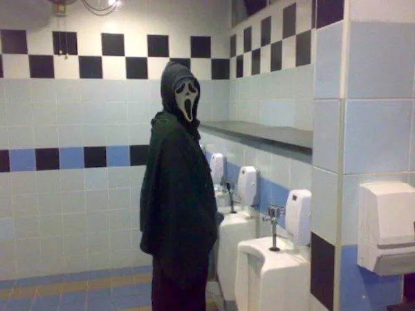 Osoba v maske strašiaka stojaca pri pisoári v kachličkovej kúpeľni.
