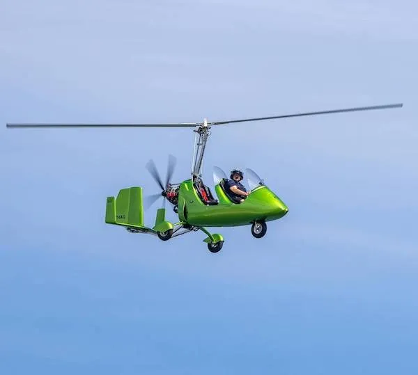 Zelený gyrokoptéra vo vzduchu s pilotom, modrá obloha na pozadí.