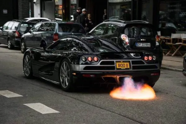 Rýchle čierne superauto vypúšťa oheň z výfuku na mestskej ulici, obklopené zaparkovanými autami.