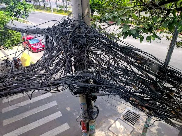 Pouličný stĺp plný zamotaných elektrických a telekomunikačných káblov pri ceste.
