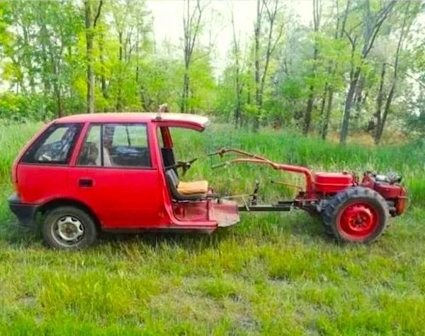 Červené auto s odstránenou prednou časťou, spojené s poľnohospodárskym traktorom, stojí na tráve pred lesom.