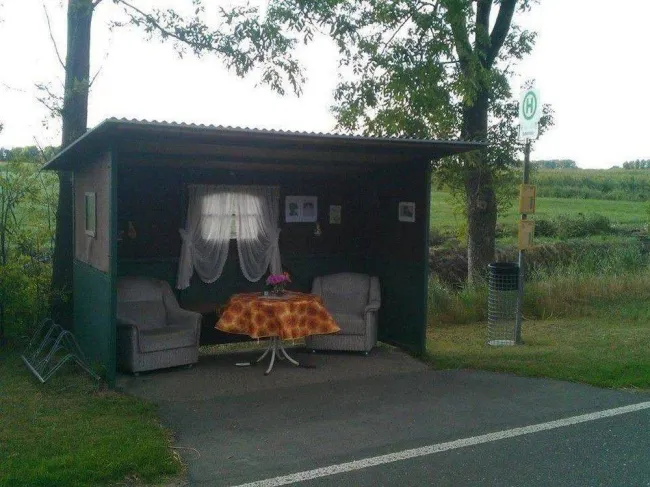 Autobusová zastávka so sedačkami a stolom, obklopená zeleňou.