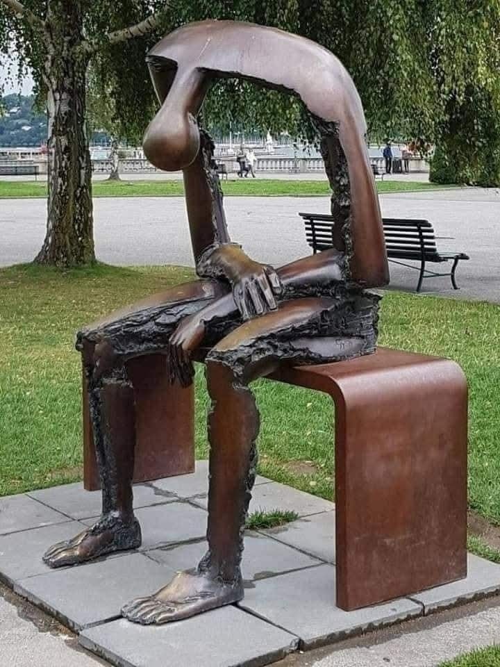 Socha sediacej postavy s ohybným chrbtom na lavičke v parku, symbolizujúca introspekciu.