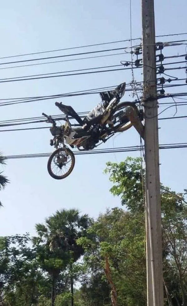 Zavesená motorka na elektrických drôtoch, pohľad zospodu, pozadie s modrým nebom a stromami.