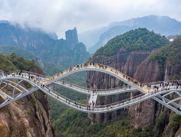Sklený most cez horskú priepasť s turistami v hmlistej horskej krajine, obklopený zeleňou a dramatickými vrchmi.