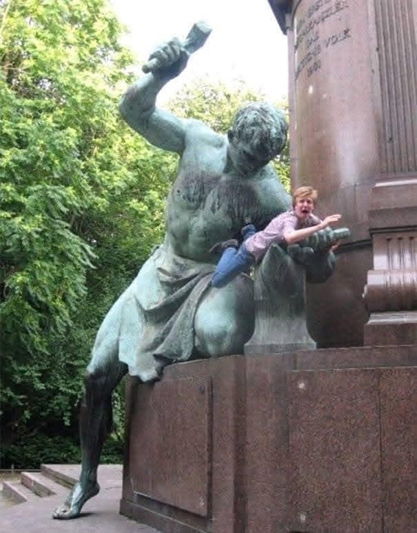 Muž ležiaci na soche s kladivom v parku, vyzerajúci prekvapene, zachytený v humornom momente.