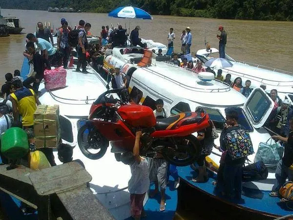Osoby na lodi prenášajú červenú motorku cez rieku, obklopené množstvom batožiny a tovaru.