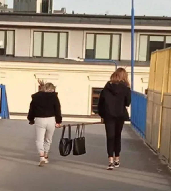 Dve osoby v tmavom oblečení kráčajú po vonkajšej chodbe, nesúc medzi sebou tašku.