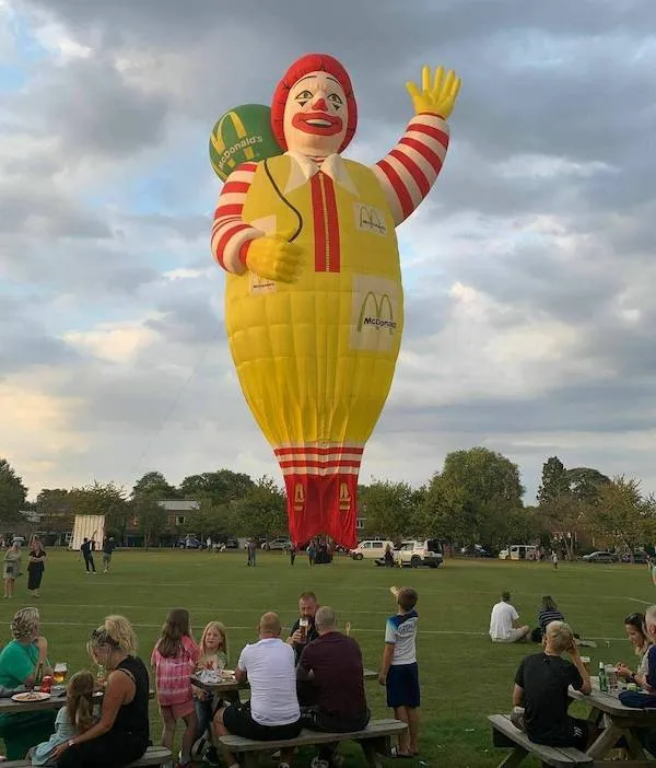 Veľký balón v tvare klauna s logom fastfoodu sa vznáša nad parkovou scénou s piknikujúcimi ľuďmi.