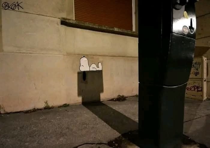 Tieň vytvára ilúziu psa Snoopyho spiaceho na boku budovy pri parkomate. Kreatívna hra svetla a tieňa.
