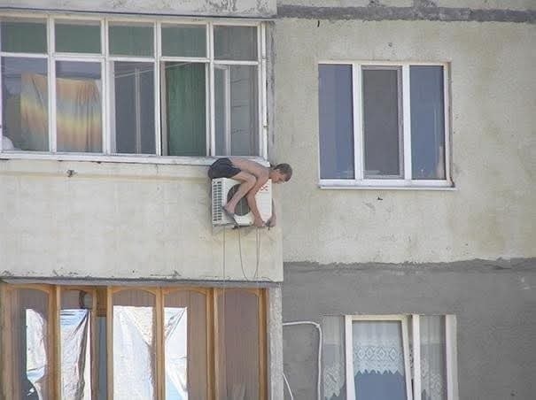 Muž v šortkách lezie po vonkajšej klimatizácii na panelákovom balkóne; bez ochrany, riskantná situácia.