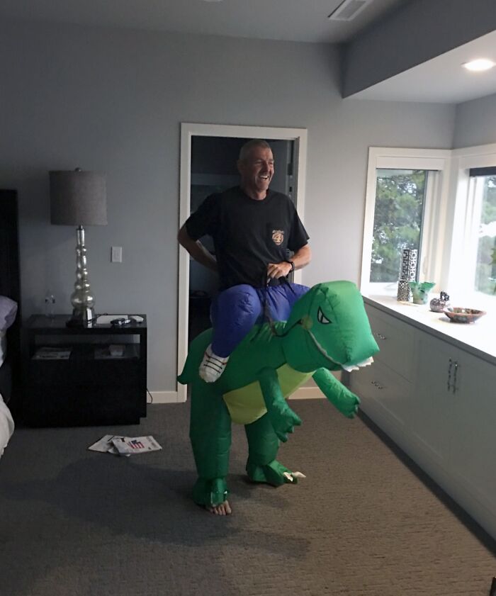 Muž v kostýme nafukovacieho dinosaura sa smeje v izbe vedľa okna s pohľadom na vonkajšok.