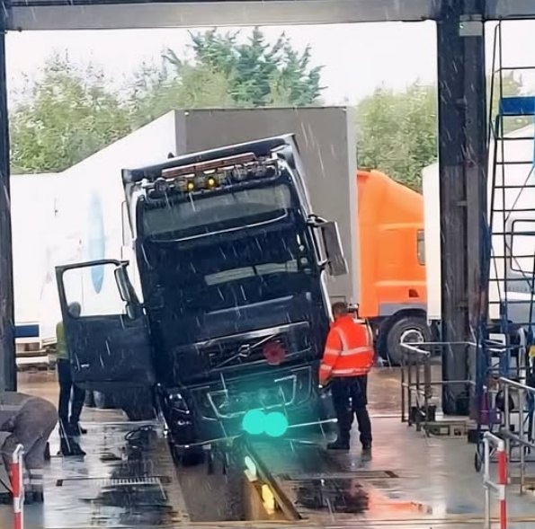 Kamión naklonený počas technickej kontroly na stanici, dvaja pracovníci ho pozorujú v daždivom počasí.