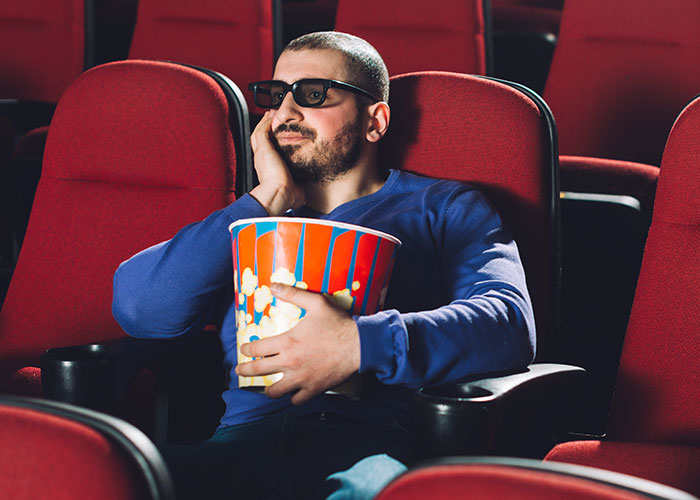 Muž v 3D okuliaroch s popcornom v kine sedí znudene na červenej sedačke.