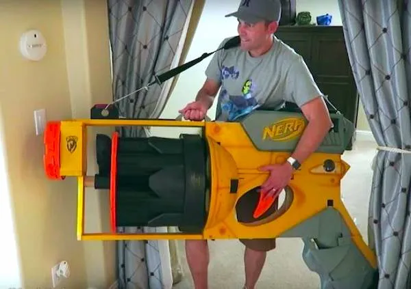 Muž nesie veľký NERF blaster v obývacej izbe, vyjadruje radosť z hračiek pre deti aj dospelých.