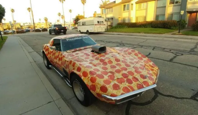 Auto s dizajnom pizze zaparkované na ulici za slnečného dňa, so zelenými palmami v pozadí.