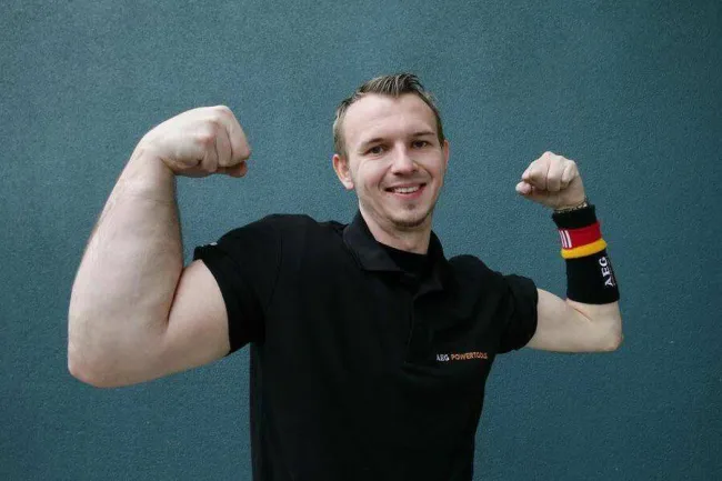 Muž v čiernom tričku pózuje s napnutými svalmi pred modrým pozadím.