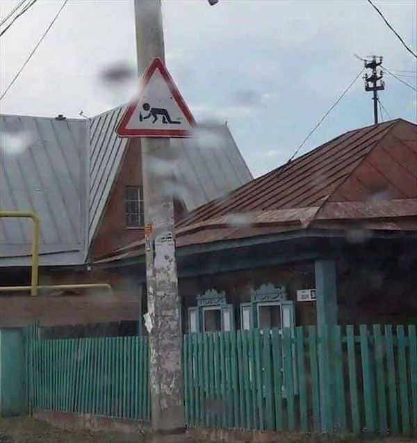 Dopravná značka so symbolom osoby na zemi pred dedinskou drevenou chatou a plotom.