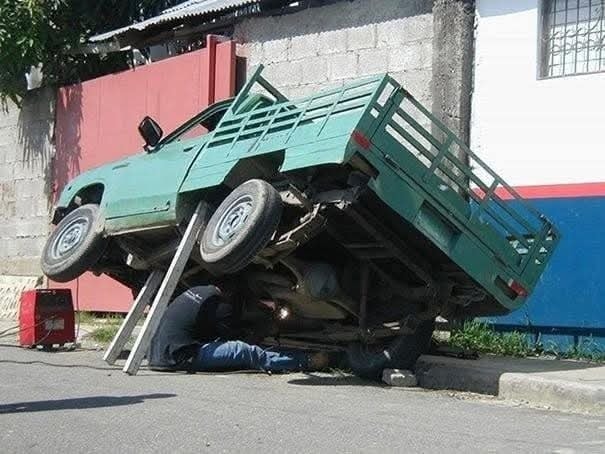 Auto zdvihnuté na ulici pre opravu podvozku, mechanik pracuje pod vozidlom. Bezpečnostné opatrenia nezabezpečené.