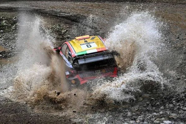 Rally auto prechádza cez štrkové blato, strieka voda; dramatická scéna športovej jazdy v teréne.