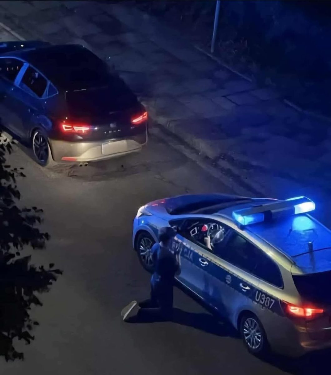 Osoba kľačí pri policajnom aute s rozsvietenými majákmi počas nočnej zastávky na ceste.