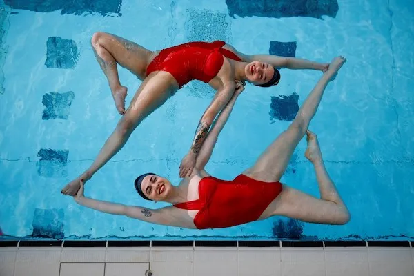 Dve ženy v červených plavkách synchronizovane plávajú v bazéne, vytvárajú geometrický vzor. Sústredenie na synchronizáciu.