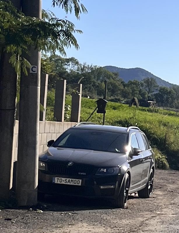 Tmavý automobil zaparkovaný na okraji cesty s výhľadom na zeleň a hory v pozadí počas slnečného dňa.