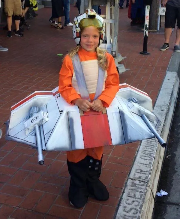 Dieťa v oranžovom kostýme pilota s lodným modelom Star Wars na festivale v meste.